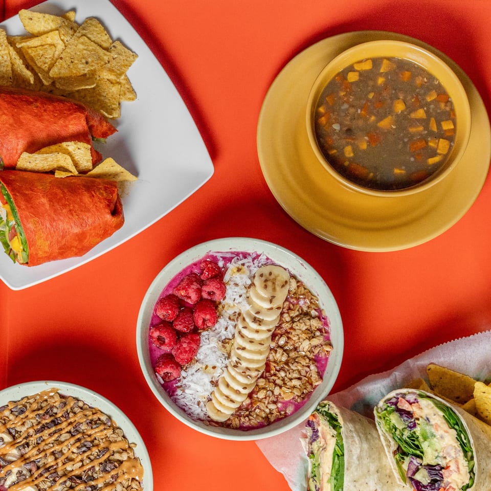 Overhead view of healthy meals including wraps, soup, and two smoothie bowls on a red background.