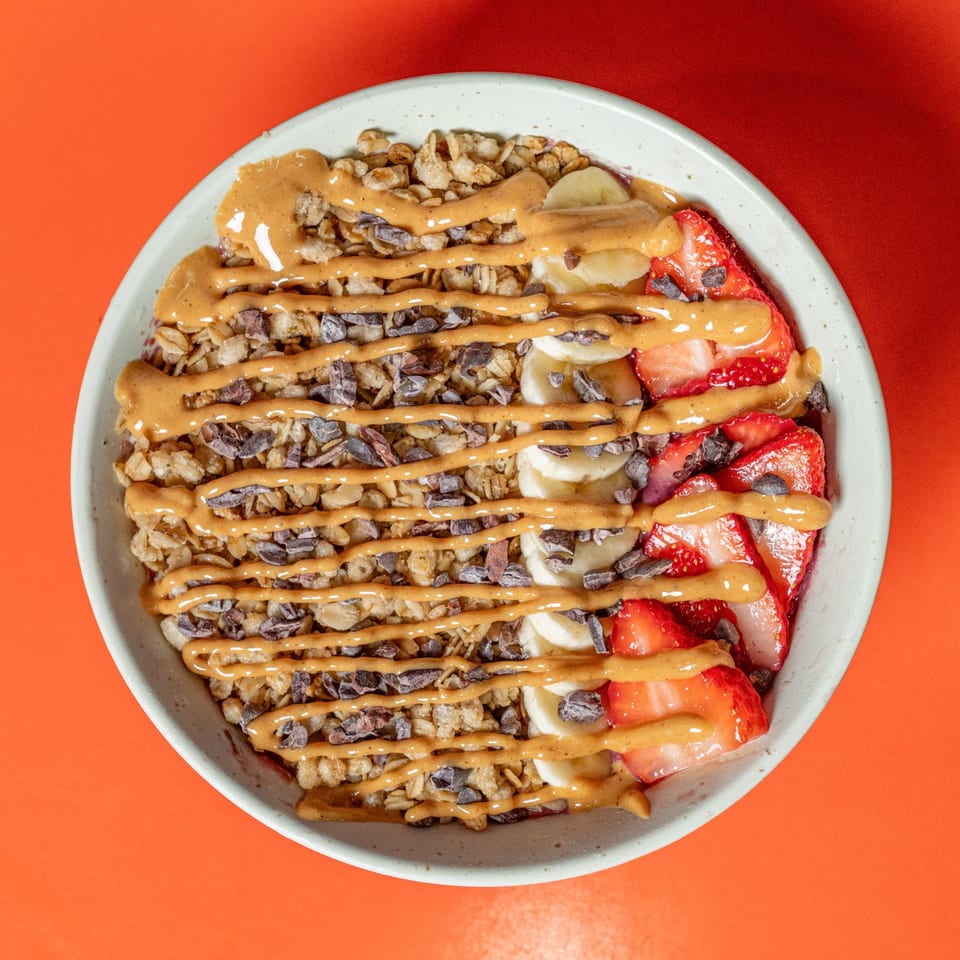 A Raw Addiction Plus acai bowl with granola, bananas, strawberries, cacao nibs, and peanut butter drizzle.