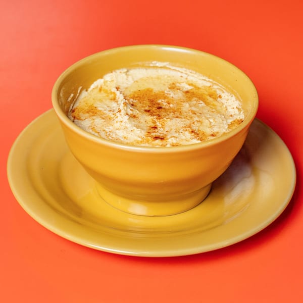 A yellow bowl of creamy hummus, topped with spices, sits on a matching saucer against a red background.